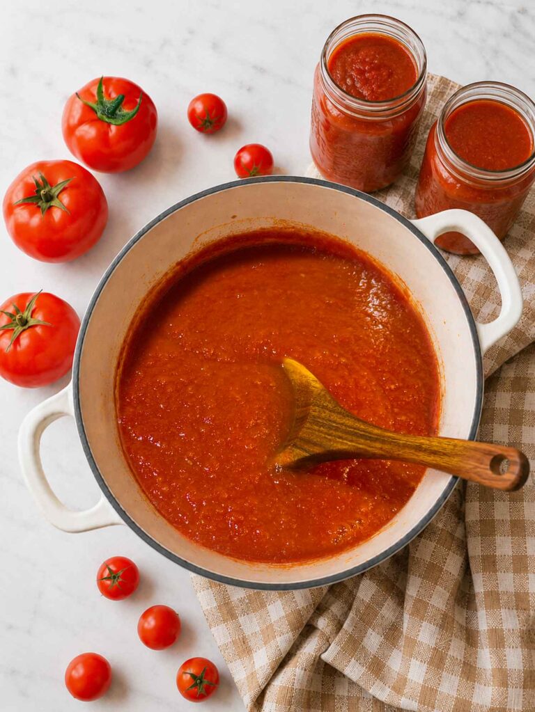 Overhead view of a pot of homemade tomato sauce with a wooden spoon, surrounded by fresh red tomatoes and two filled canning jars on a white quartz counter
