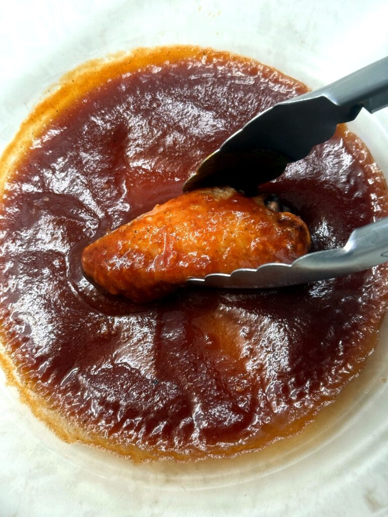 A crispy wing being turned in a bowl of homemade BBQ sauce with tongs
