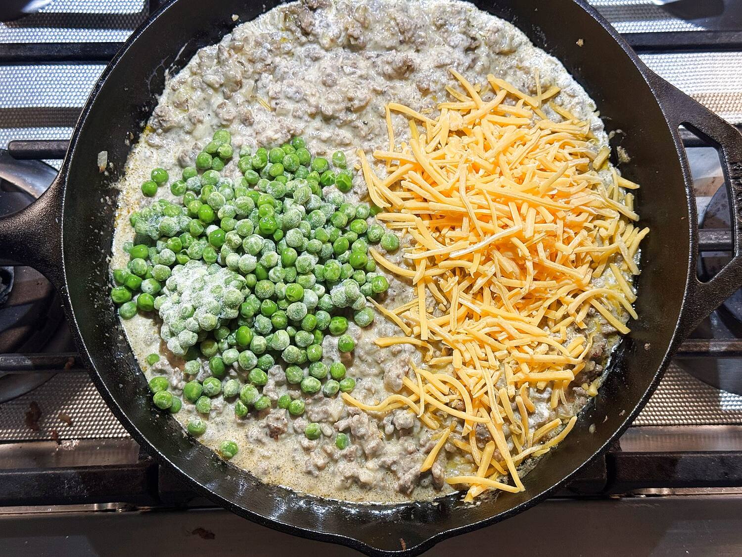 Frozen peas and shredded sharp cheddar added to the creamy beef filling in a cast iron skillet, before stirring.