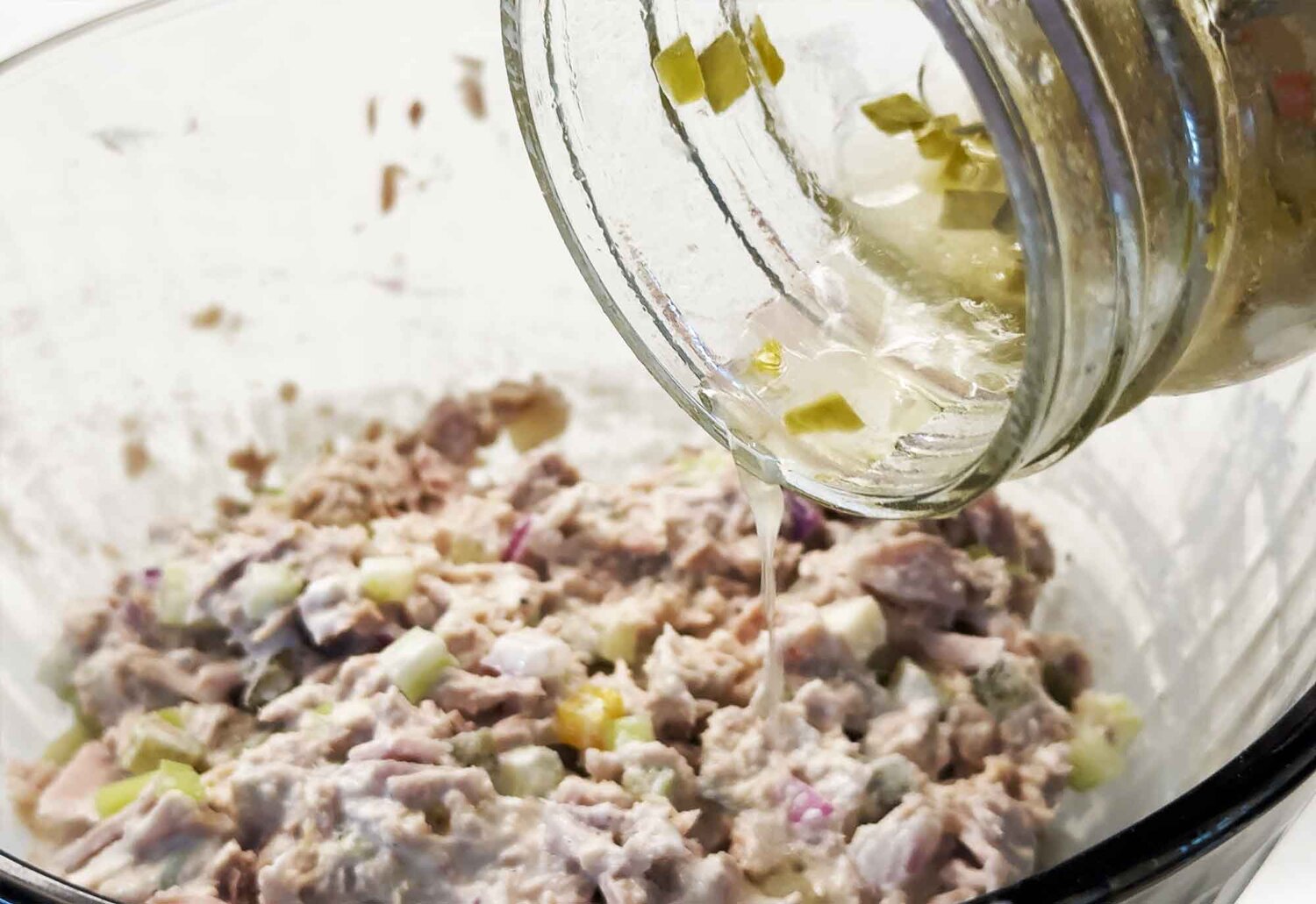 Pouring a splash of sweet relish brine from a jar into a bowl of tuna salad