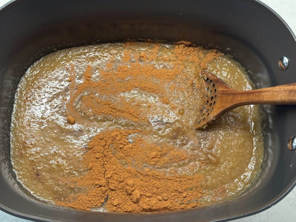 Ground cinnamon and warm spices sprinkled over smooth apple pulp in a slow cooker, being stirred with a wooden spoon