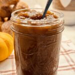 An open jar of apple butter with a spoon, next to a plate of cinnamon sugar apple cider donuts and a small pumpkin