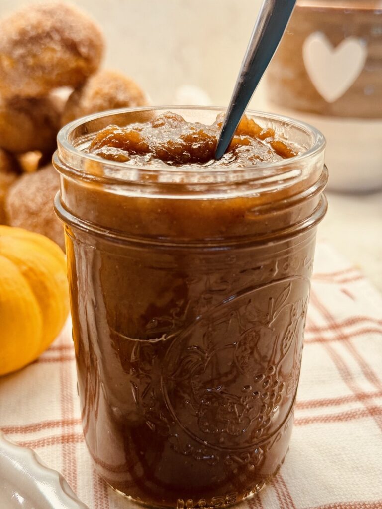 An open jar of apple butter with a spoon, next to a plate of cinnamon sugar apple cider donuts and a small pumpkin