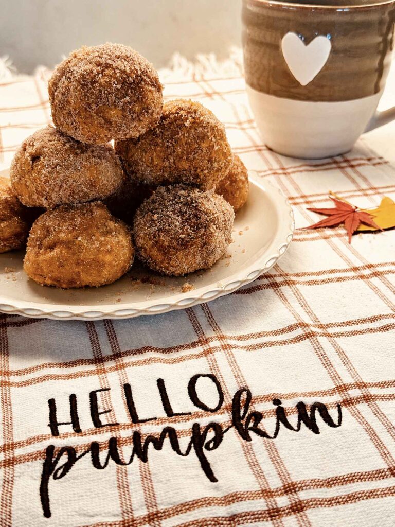 Baked pumpkin doughnut holes coated in cinnamon sugar, stacked on a white plate beside a mug of coffee