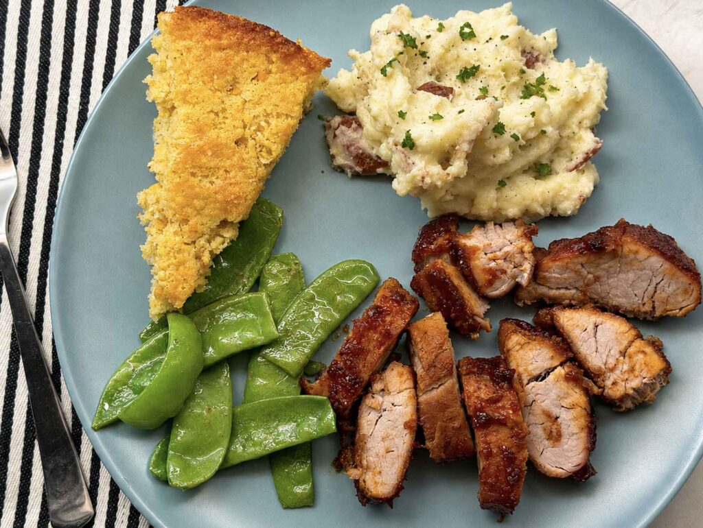 A dinner plate of sliced BBQ pork tenderloin with mashed potatoes, snap peas, and cornbread
