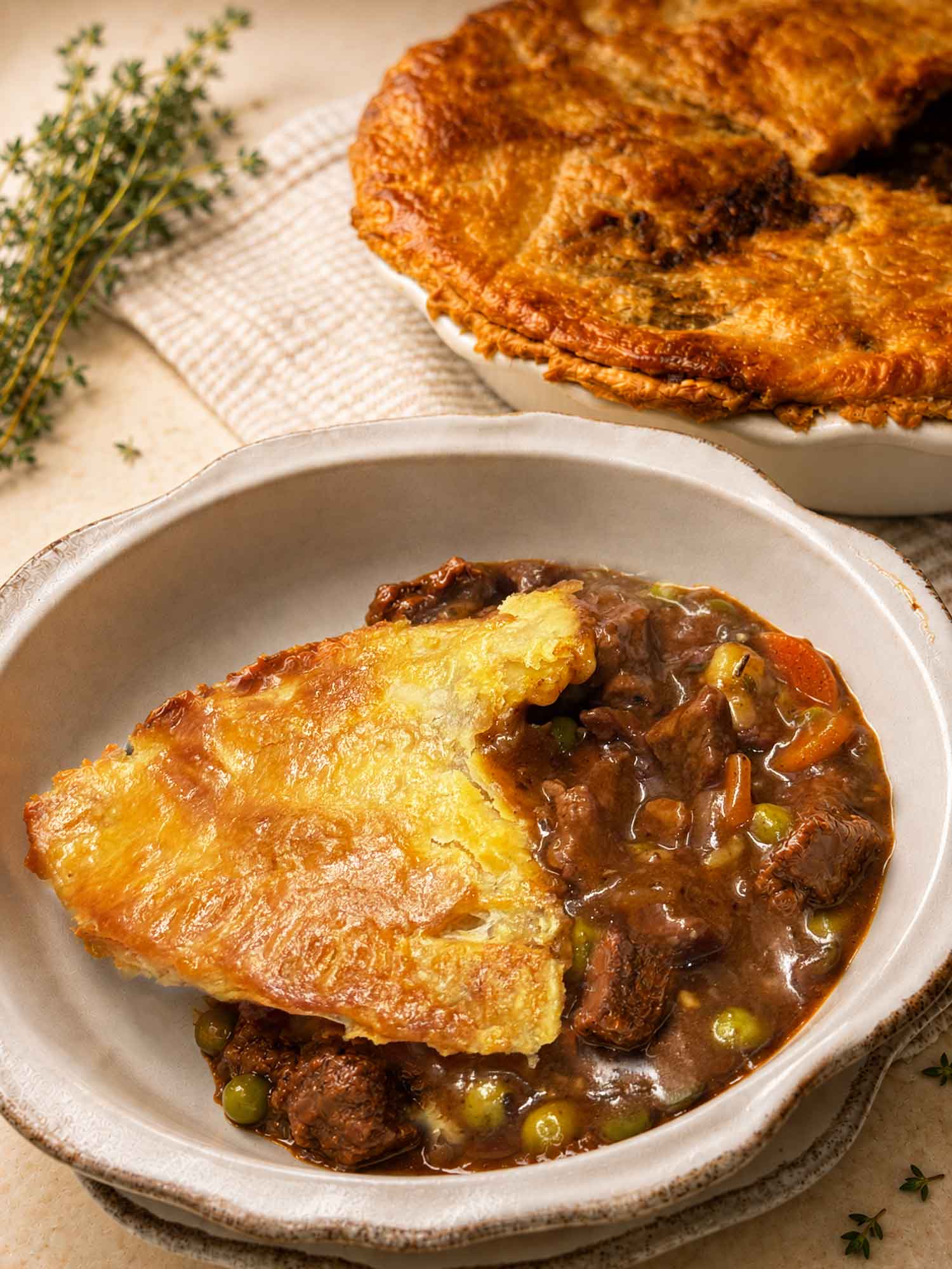 A bowl of beef pot pie with flaky crust, tender beef, peas, and gravy, with the pie dish behind