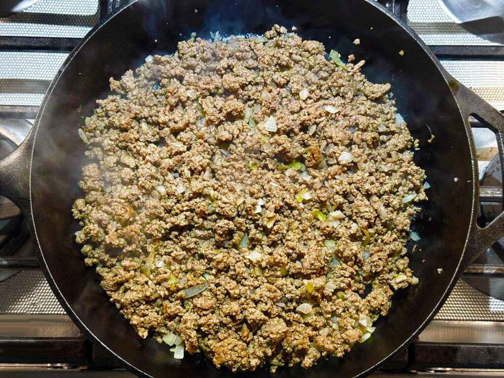Ground beef and diced onion browned together in a cast iron skillet on the stove.