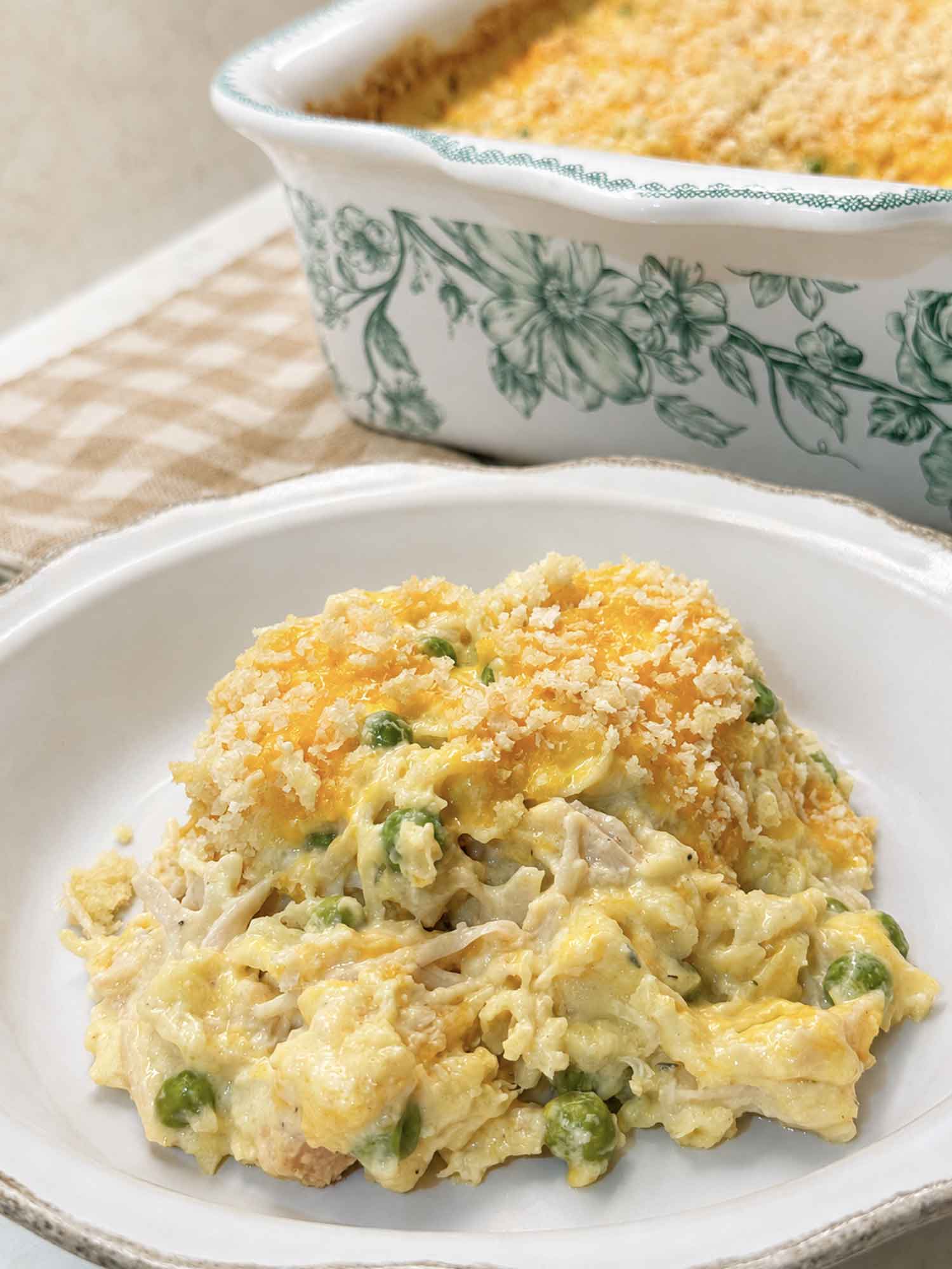 A bowl of creamy chicken noodle casserole with peas and a golden panko topping, served in front of the baking dish