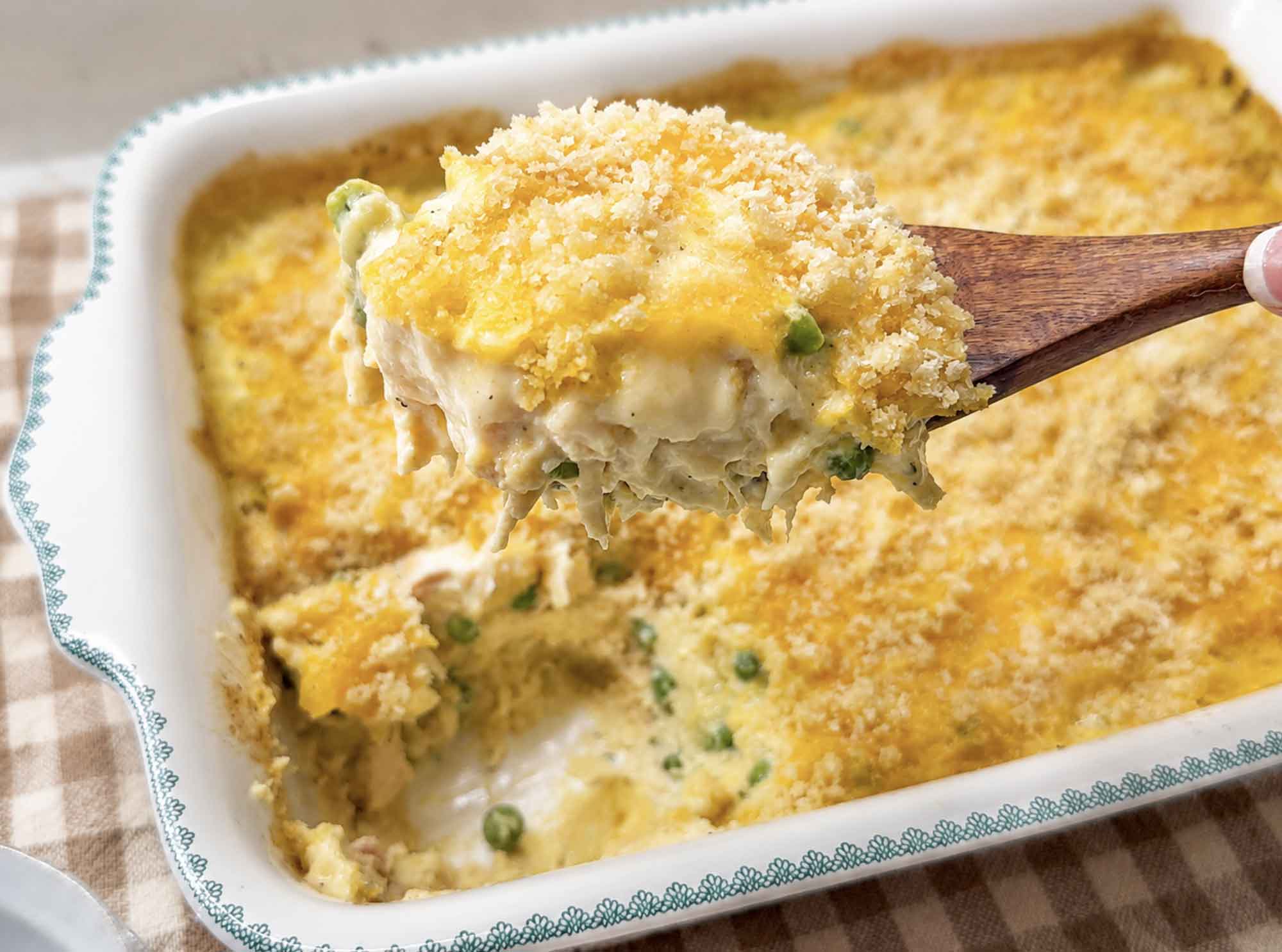 A wooden spoon lifting a serving of creamy chicken noodle casserole with peas from the baking dish