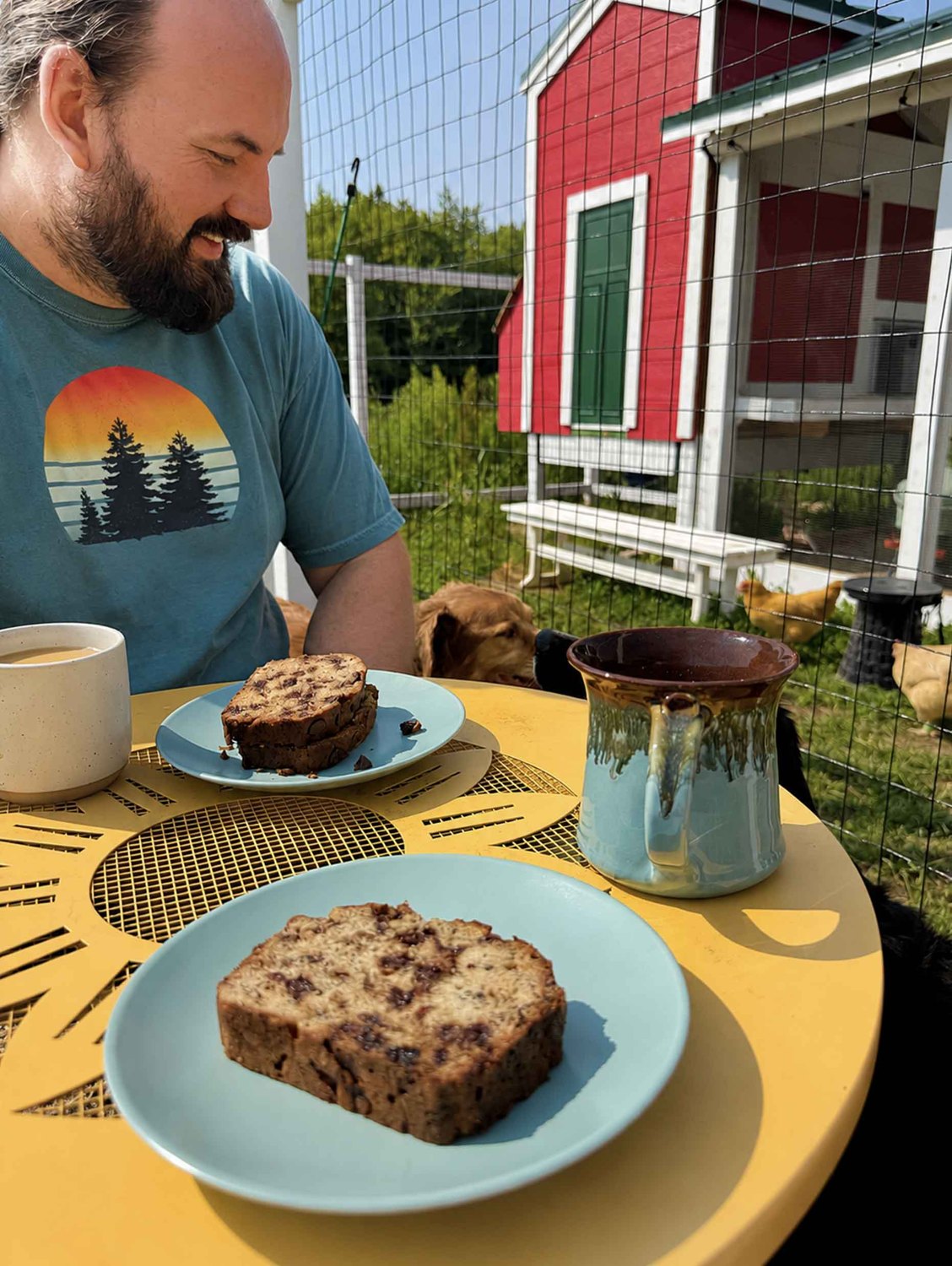 Greg enjoying a slice of chocolate chip banana bread with a cup of coffee at a table outside by the red barn