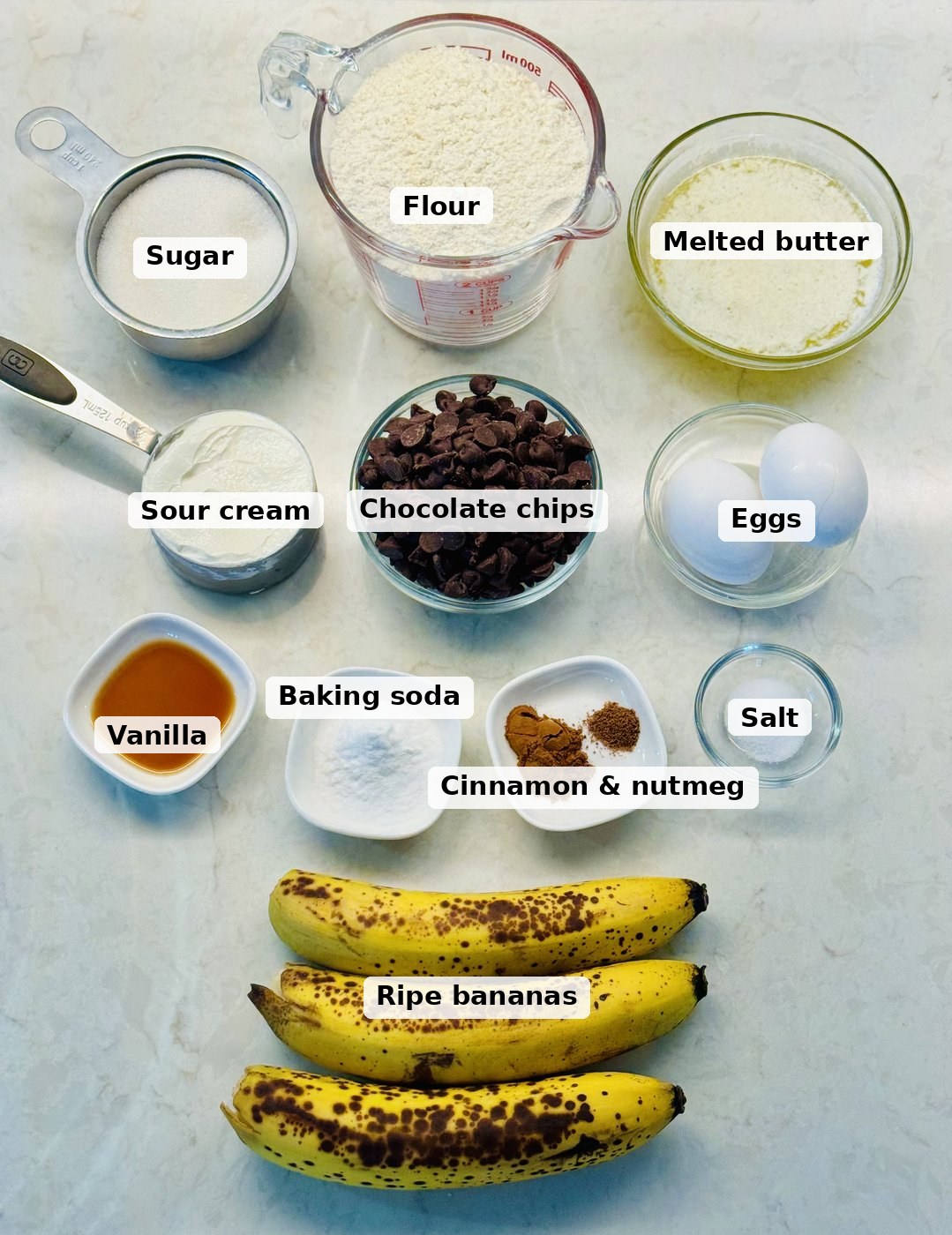 Labeled ingredients for chocolate chip banana bread: ripe bananas, flour, sugar, melted butter, sour cream, eggs, vanilla, baking soda, cinnamon and nutmeg, salt, and semi-sweet chocolate chips