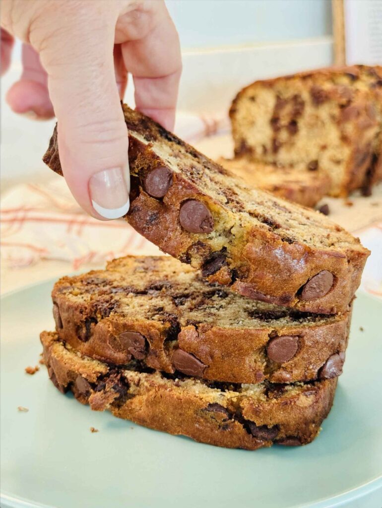 A hand lifting a slice of chocolate chip banana bread from a stack on a plate, showing the soft chocolate-studded interior