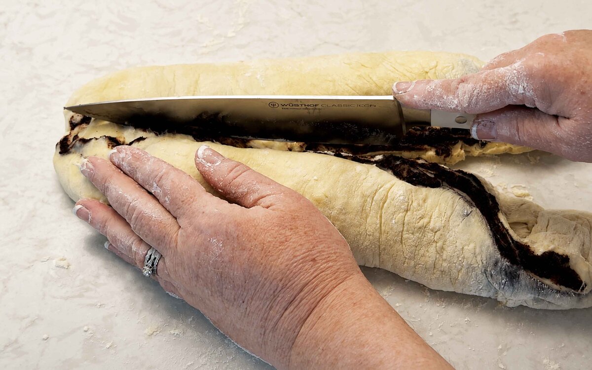 Cutting the rolled babka log in half lengthwise with a knife to expose the chocolate layers