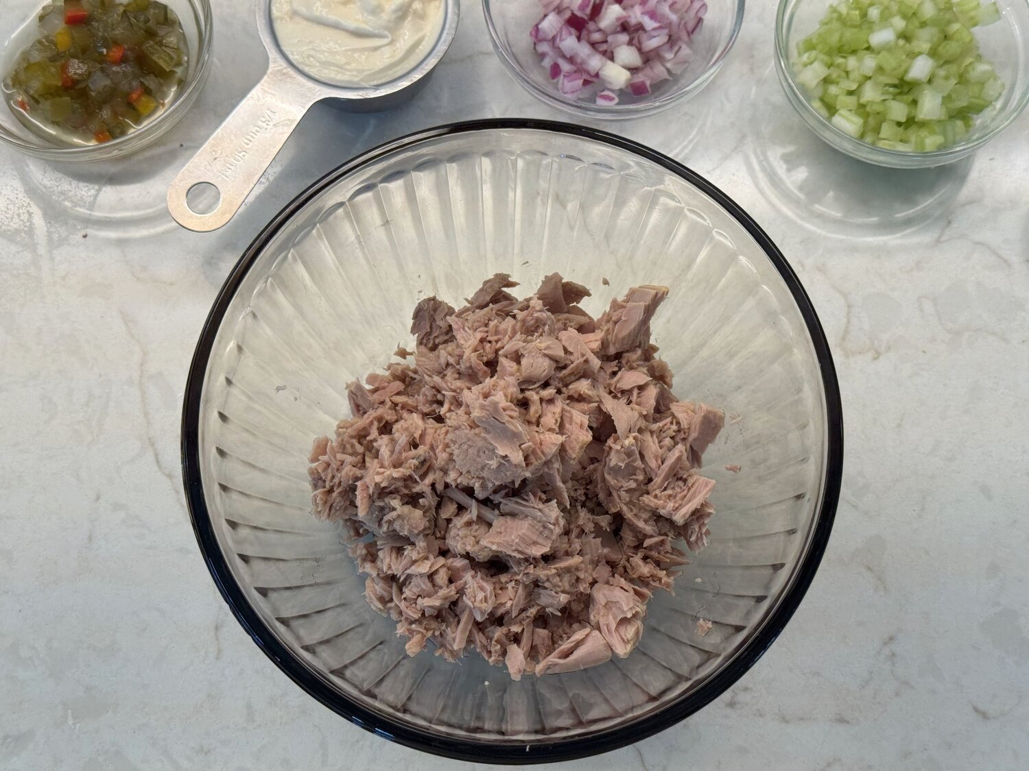 Well-drained tuna flaked in a glass bowl next to the other tuna salad ingredients
