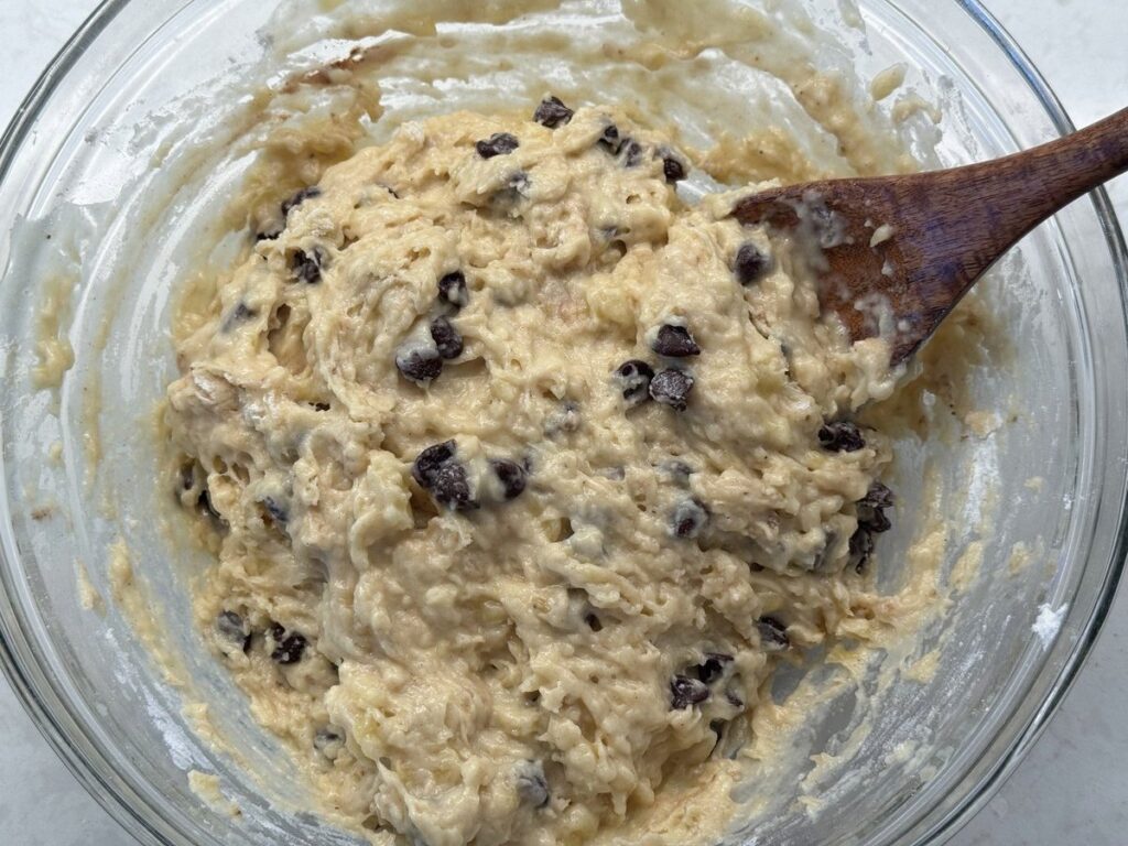 Chocolate chips folded into thick banana bread batter with a wooden spoon