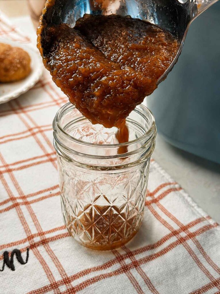 Ladling hot apple butter into a quilted half-pint canning jar on a plaid kitchen towel