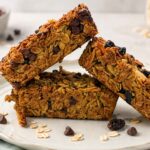 A stack of homemade oatmeal breakfast bars on a plate, chocolate chip and cherry-almond, with oats and chocolate chips scattered around.