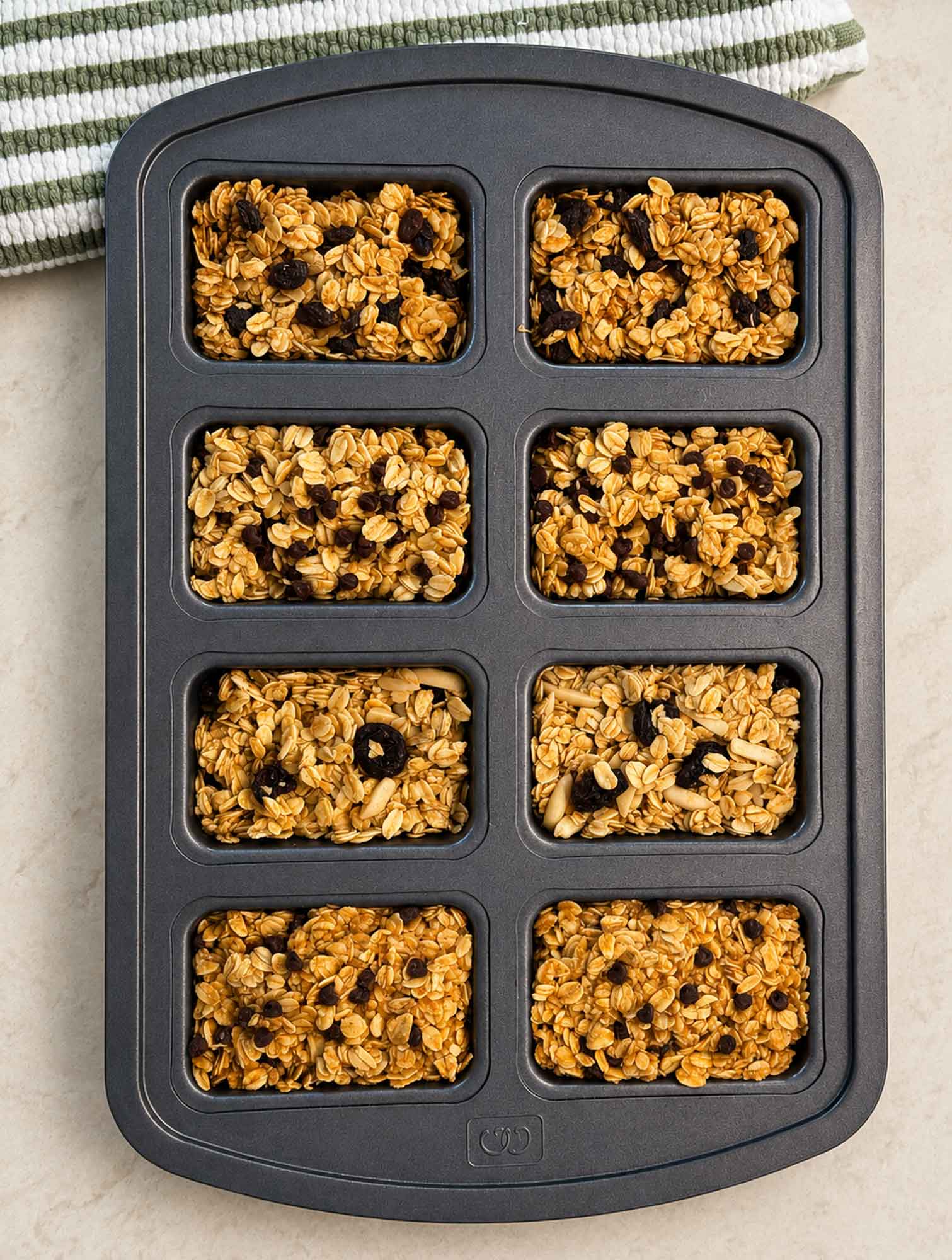 A mini loaf pan with eight cavities filled with different flavors of raw oatmeal bar mixture, pressed down and ready to bake.