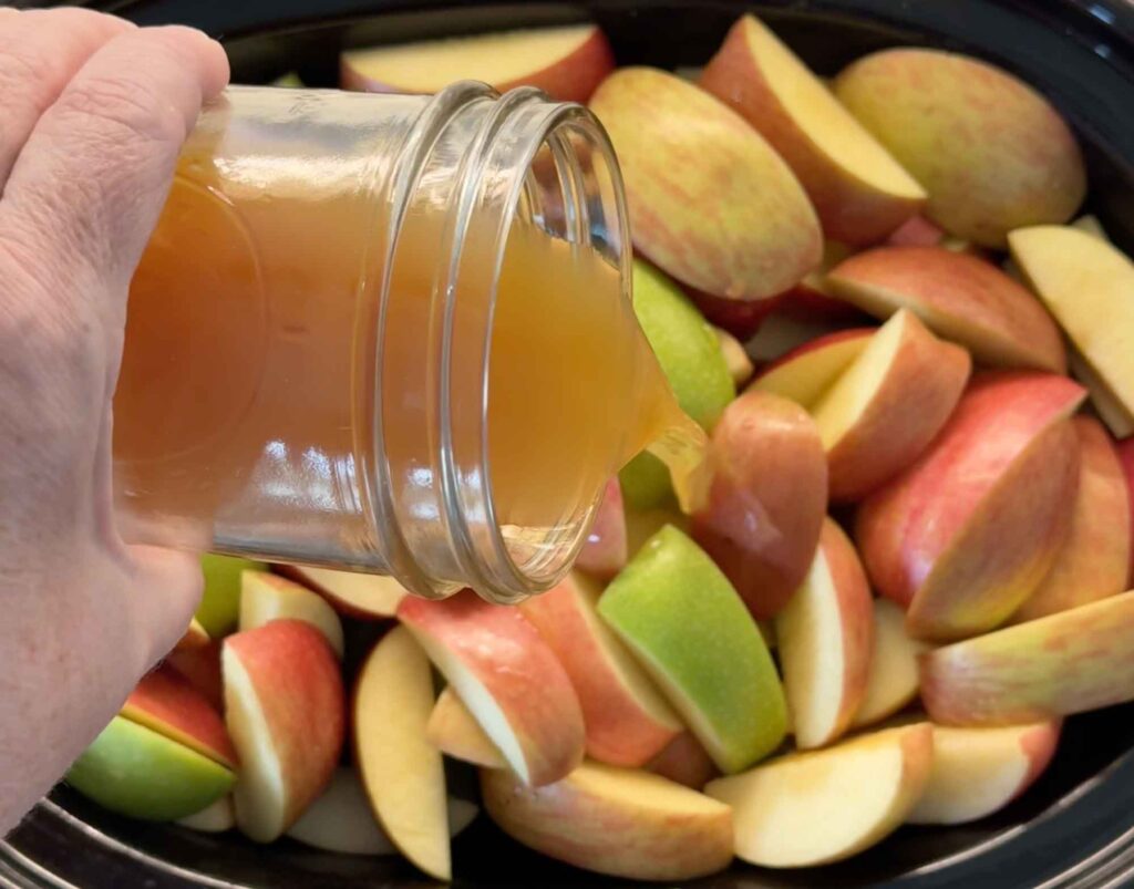 Pouring sweet apple cider from a glass jar over quartered apples in a slow cooker