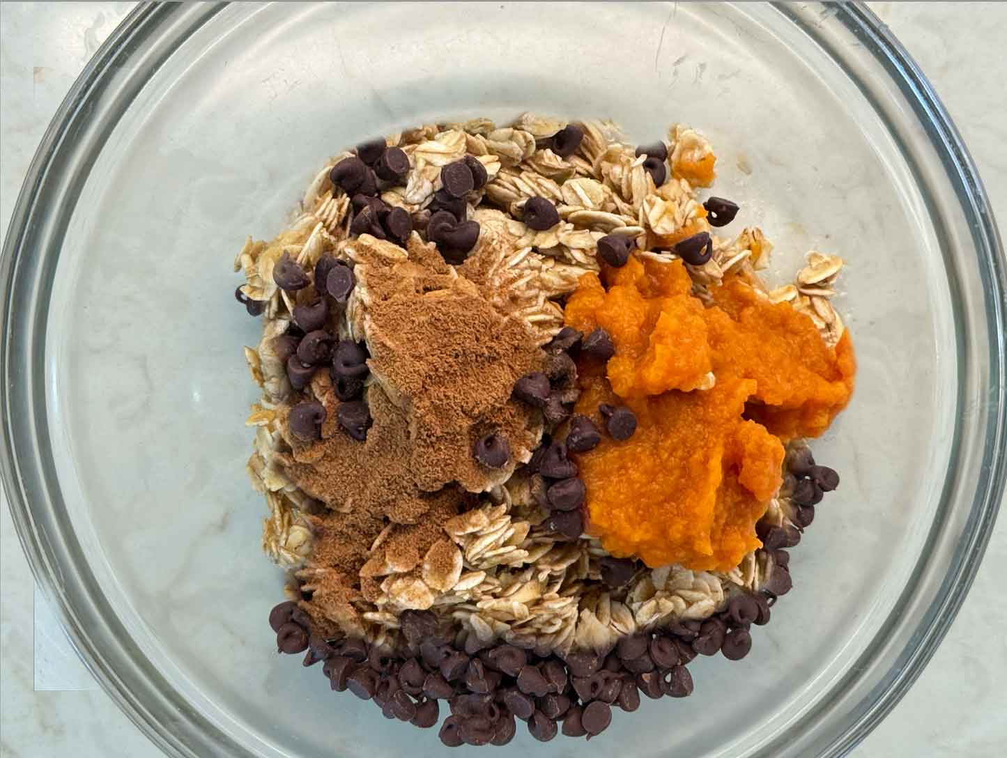 A glass bowl of oat mixture topped with pumpkin puree, pumpkin pie spice, and chocolate chips, ready to stir together.