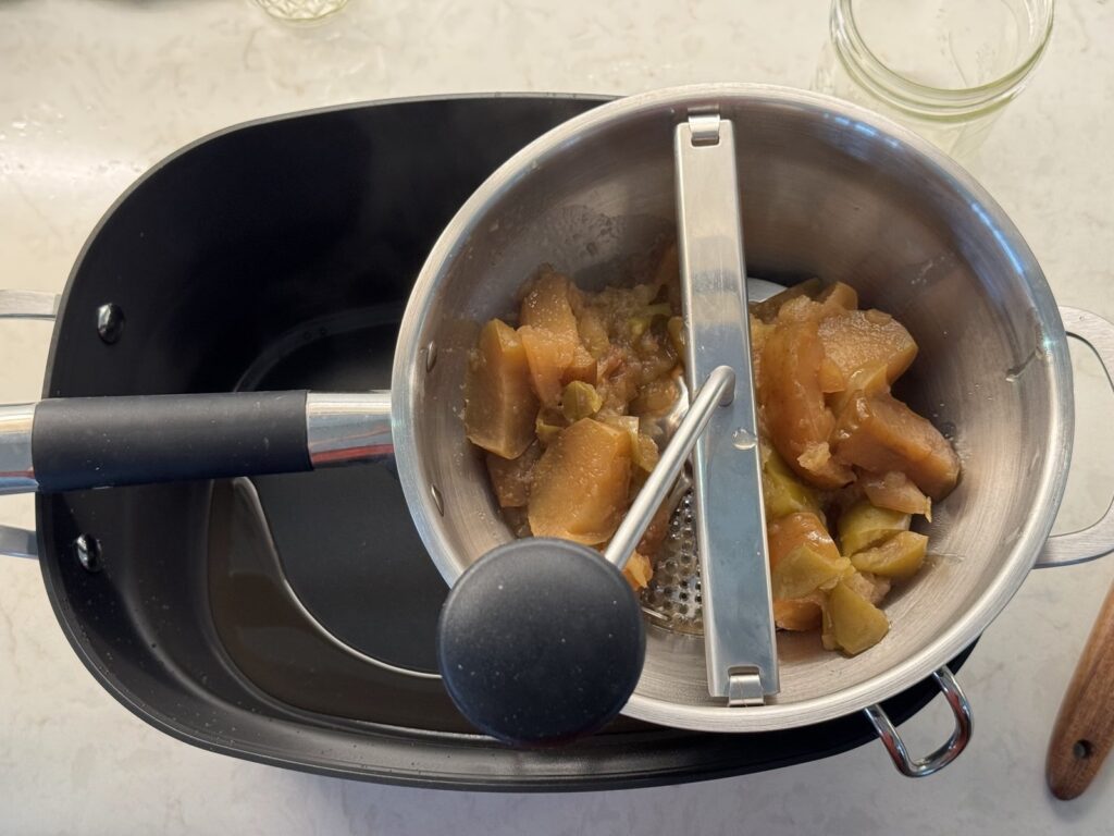Cooked apples in a food mill set over a stainless steel pot, with the peels separating out