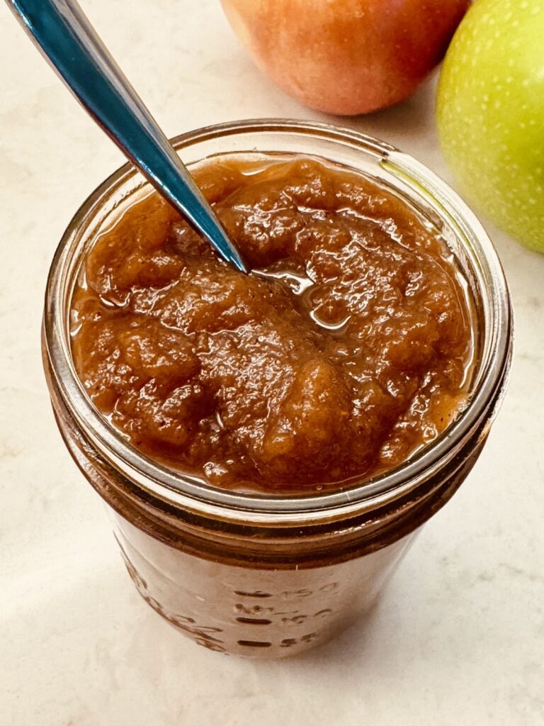 A half-pint jar of thick homemade slow cooker apple butter with a spoon in it, with a red and a green apple behind it