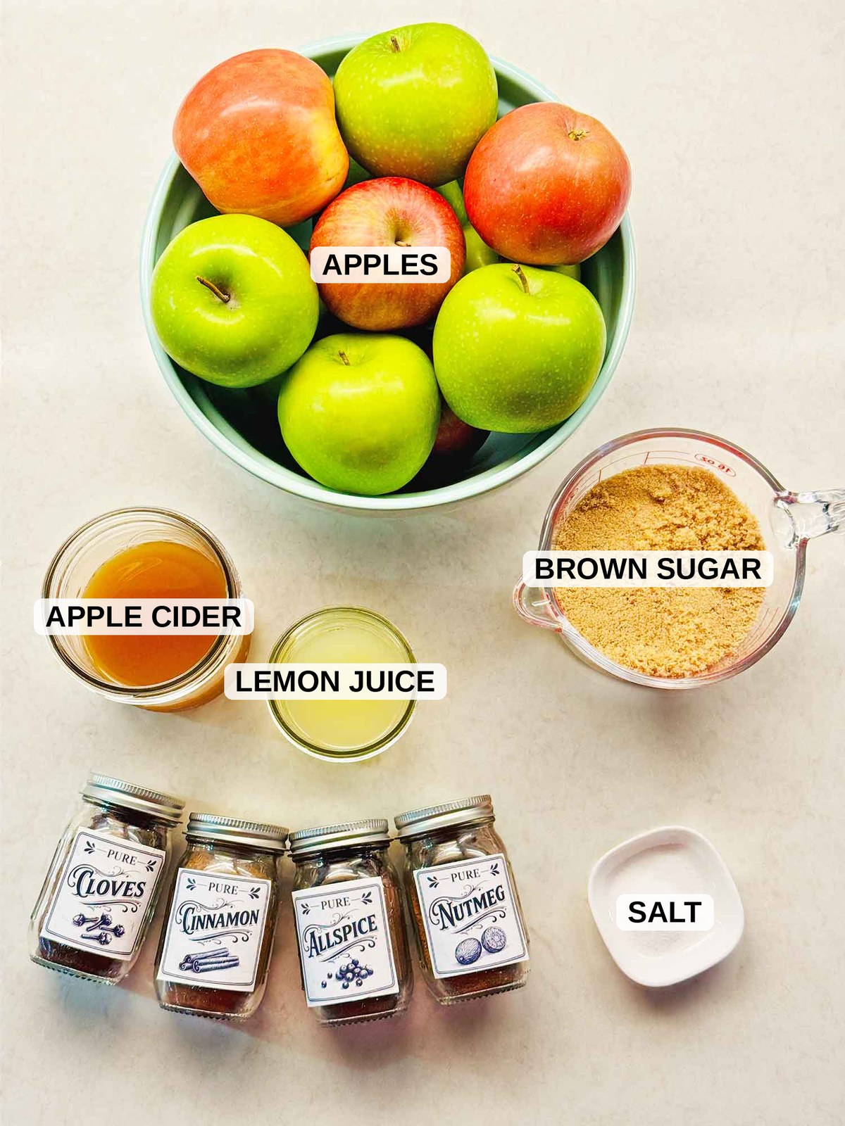 Ingredients for slow cooker apple butter labeled on a counter: apples, apple cider, lemon juice, brown sugar, cinnamon, cloves, allspice, nutmeg, and salt