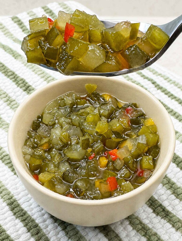 A spoonful of homemade sweet pickle relish held above a bowl, showing chunky cucumbers and pepper flecks