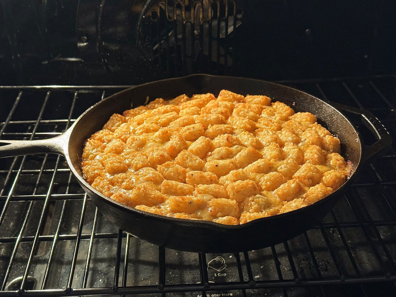 Tater tot casserole baking in a cast iron skillet on an oven rack, the tots turned golden brown.