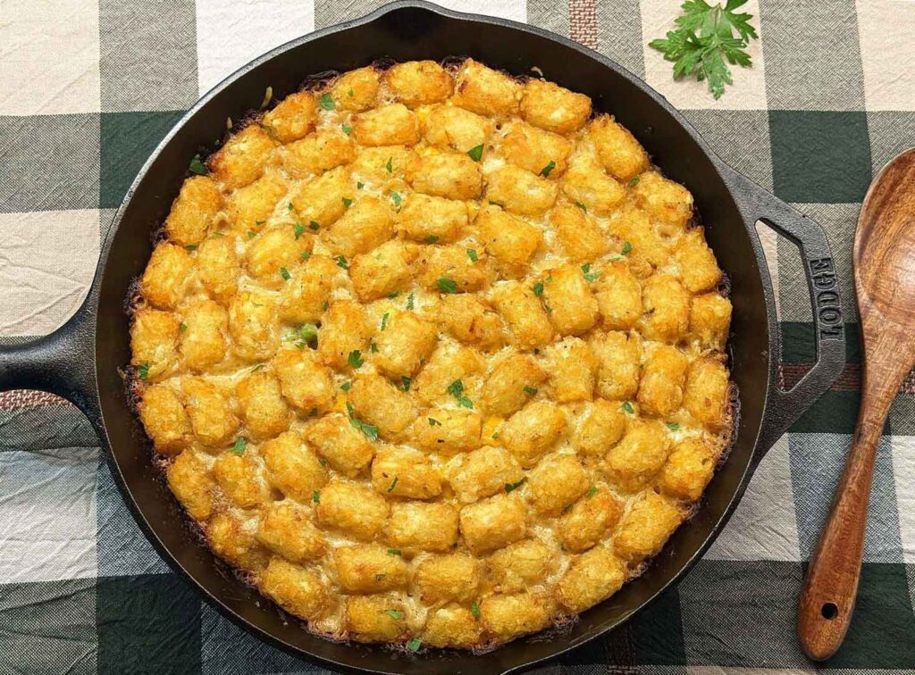 Baked tater tot casserole in a cast iron skillet on a green checked tablecloth, with a wooden spoon and a sprig of parsley alongside.