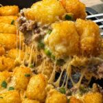 Tater Tot Casserole (One-Pan Comfort Food) Serving cheesy tater tot casserole lifted from the cast iron skillet, showing cheesy pulls from the creamy ground beef, peas, and melted cheddar under the tater tots.