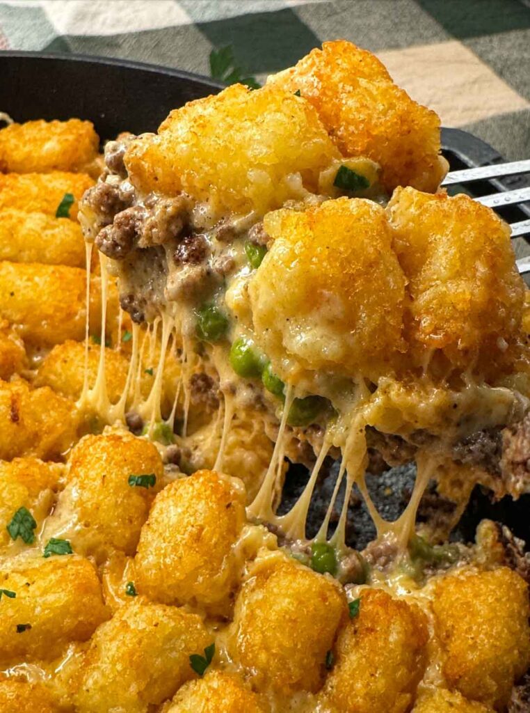 Serving cheesy tater tot casserole lifted from the cast iron skillet, showing cheesy pulls from the creamy ground beef, peas, and melted cheddar under the tater tots.