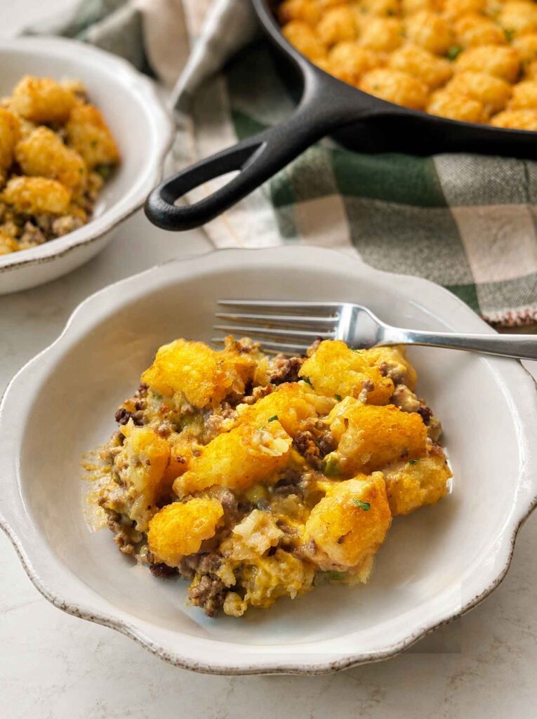 A serving of tater tot casserole in a white bowl with a fork, the cast iron skillet and a second bowl in the background.