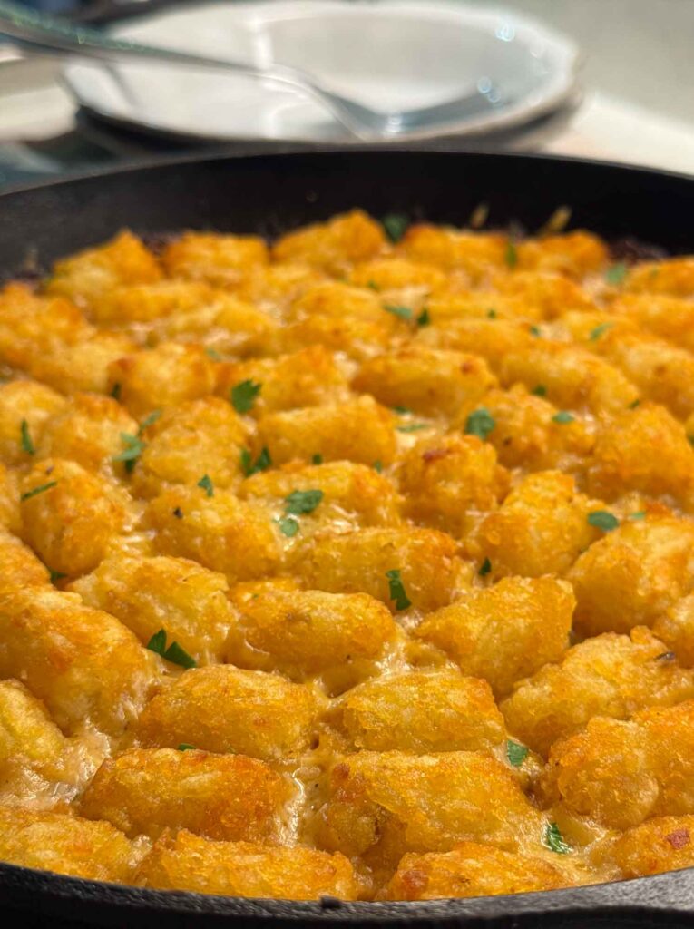 Tater tot casserole baked in a cast iron skillet, the tater tots crisp and golden and scattered with chopped parsley, with stacked plates and a serving spoon behind.
