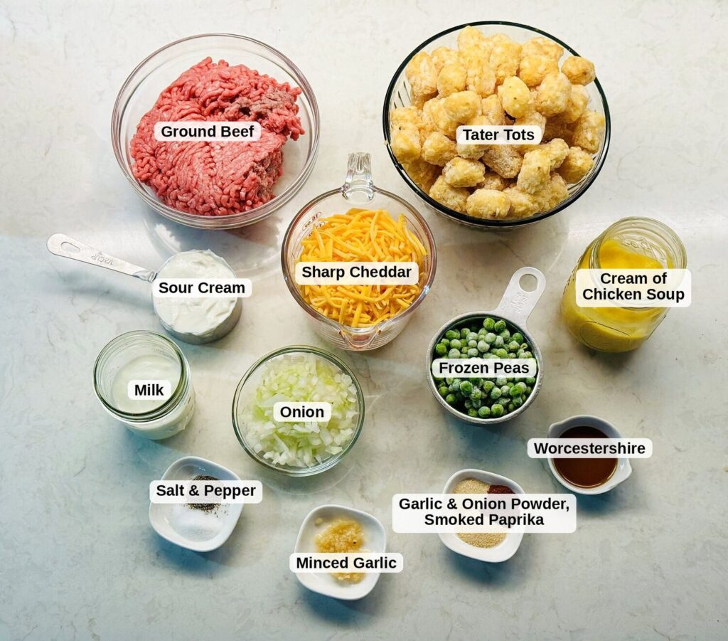 Labeled ingredients for tater tot casserole: ground beef, tater tots, sharp cheddar, sour cream, cream of chicken soup, milk, onion, frozen peas, Worcestershire sauce, minced garlic, salt and pepper, garlic and onion powder, and smoked paprika.