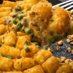 A spoonful of tater tot casserole lifted from the cast iron skillet, showing creamy ground beef, peas, and melted cheddar under the crisp tater tots.