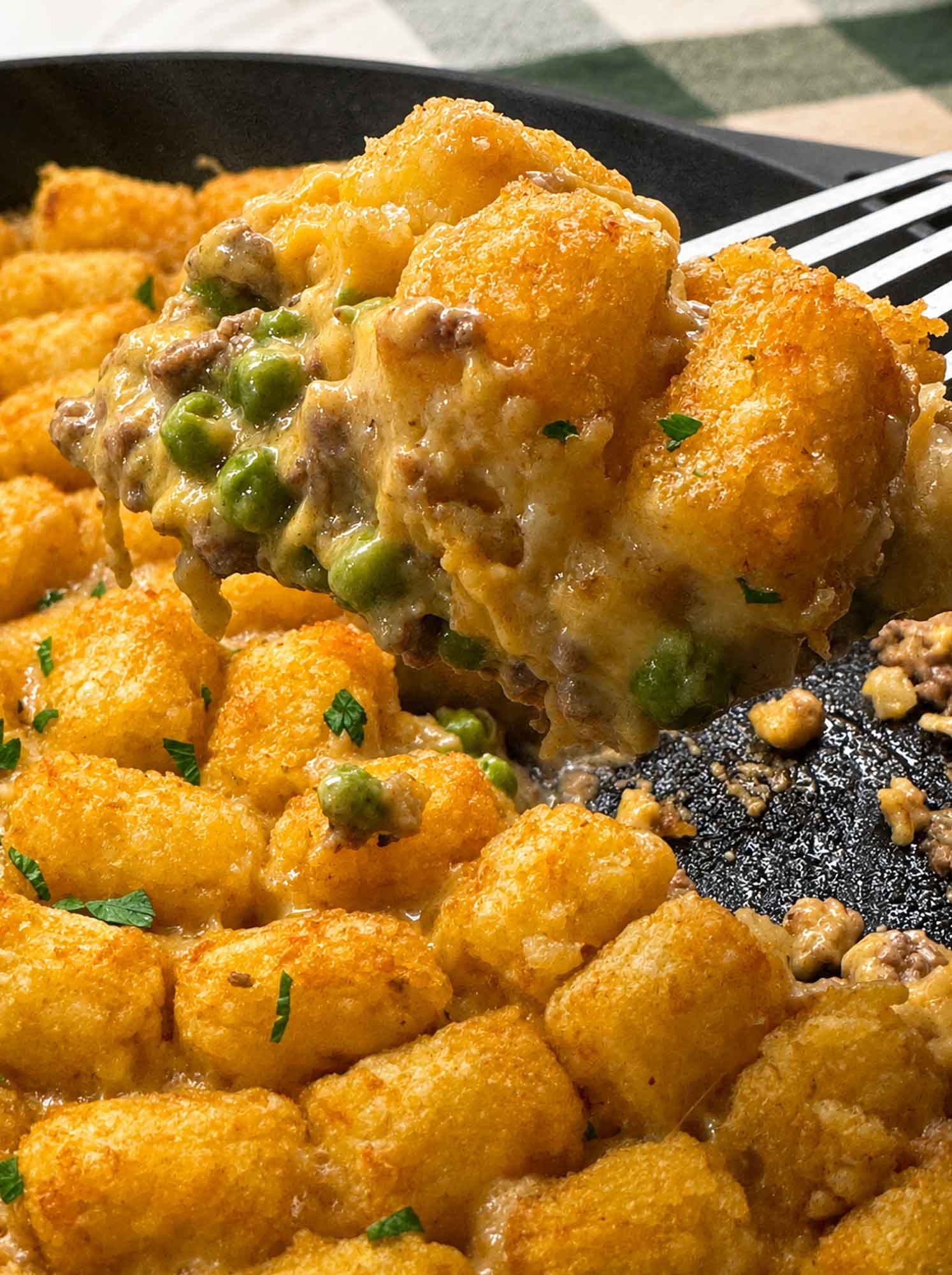 A spoonful of tater tot casserole lifted from the cast iron skillet, showing creamy ground beef, peas, and melted cheddar under the crisp tater tots.