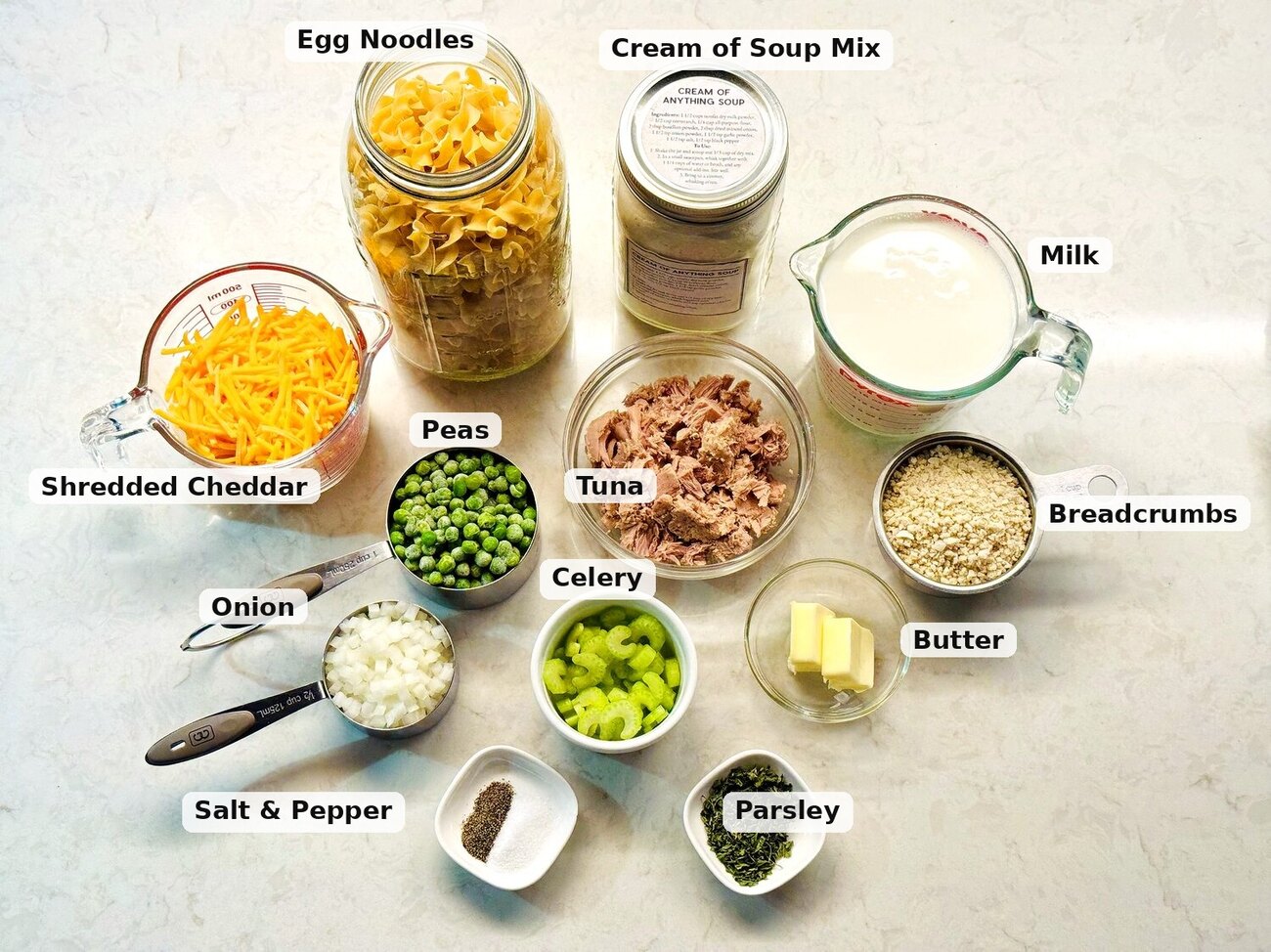 Tuna noodle casserole ingredients laid out: egg noodles, tuna, peas, cheddar, onion, celery, butter, and breadcrumbs