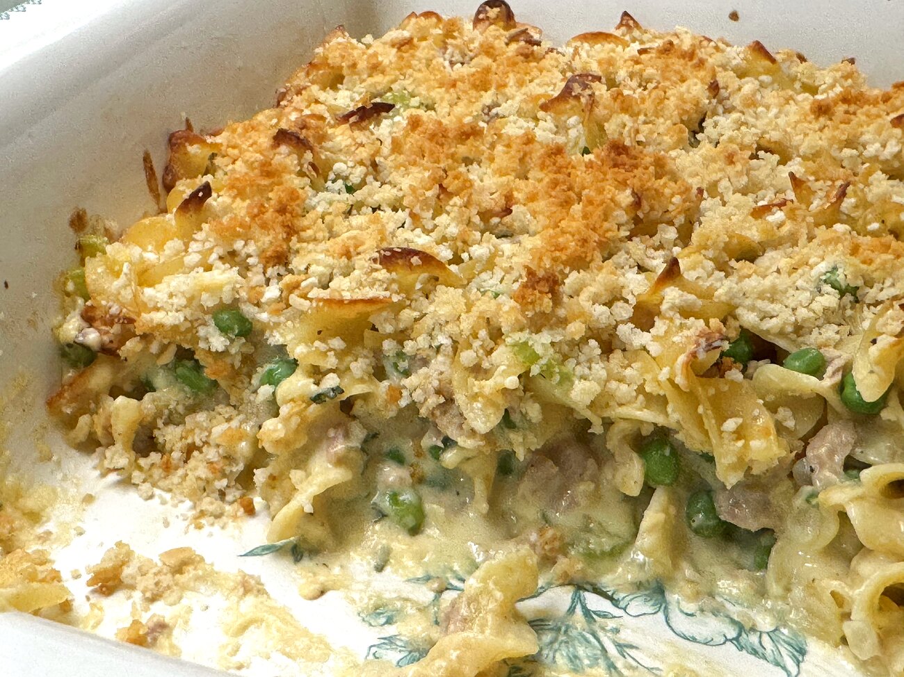 A serving spoon lifting creamy tuna noodle casserole from the dish, showing peas and noodles inside