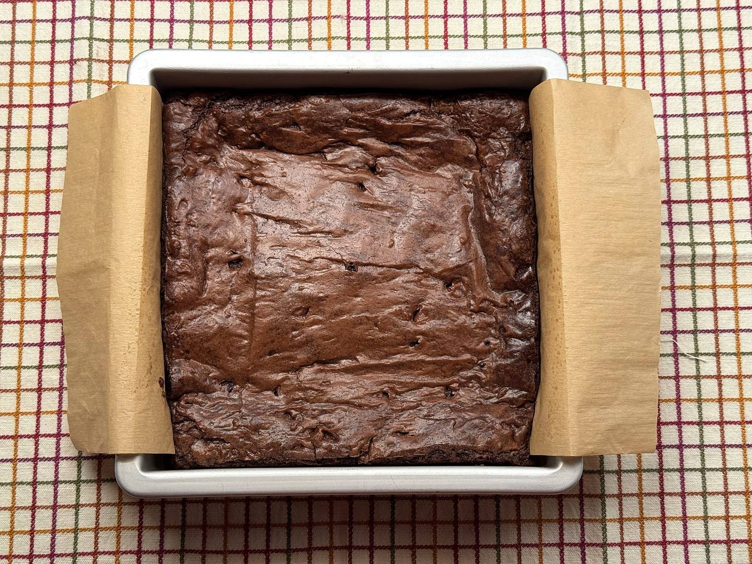 Overhead view of a pan of baked brownies with a shiny, rippled top in a parchment-lined 8x8 pan on a plaid towel