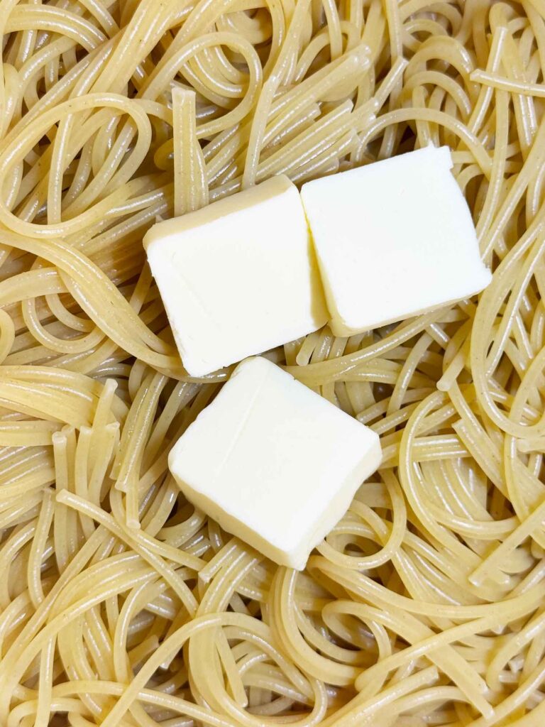 Three pats of butter melting into a pot of hot drained spaghetti for million dollar spaghetti.