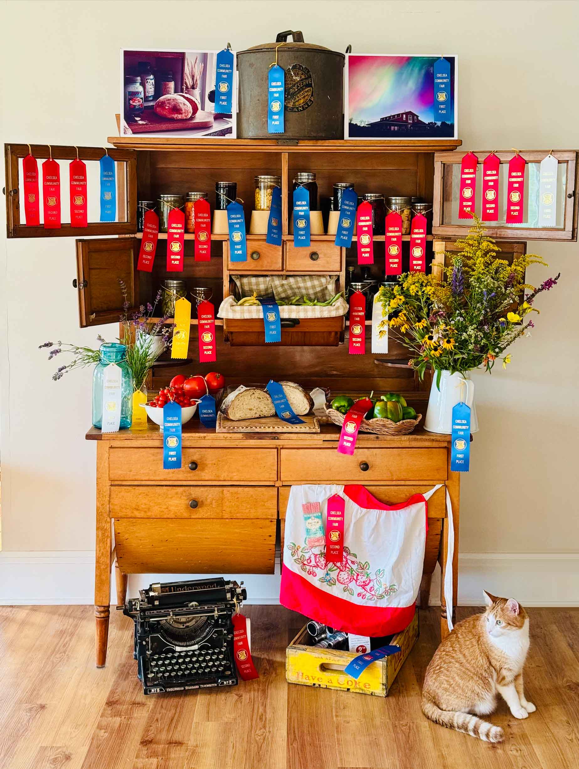 Antique Hoosier cabinet covered in Chelsea Community Fair ribbons with home canned jars, sourdough bread, tomatoes, and wildflowers.