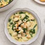 How to Make Chicken Gnocchi Soup (Better Than Olive Garden) A scalloped white bowl of creamy chicken gnocchi soup with potato gnocchi, diced chicken breast, spinach, and matchstick carrots, with breadsticks and the Dutch oven alongside.
