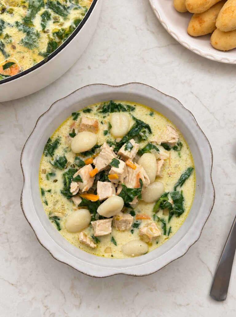 A scalloped white bowl of creamy chicken gnocchi soup with potato gnocchi, diced chicken breast, spinach, and matchstick carrots, with breadsticks and the Dutch oven alongside.