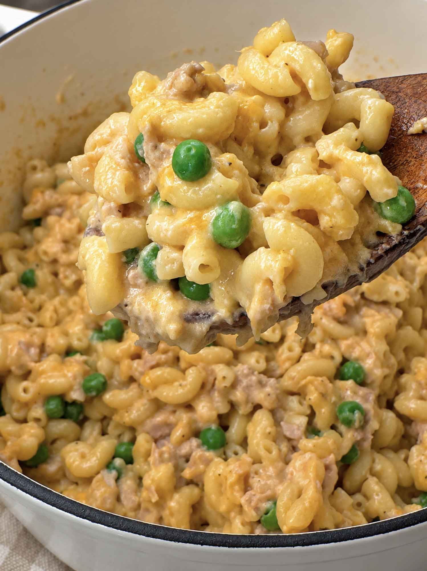 A wooden spoon lifting a serving of creamy tuna pasta with visible chunks of tuna and peas