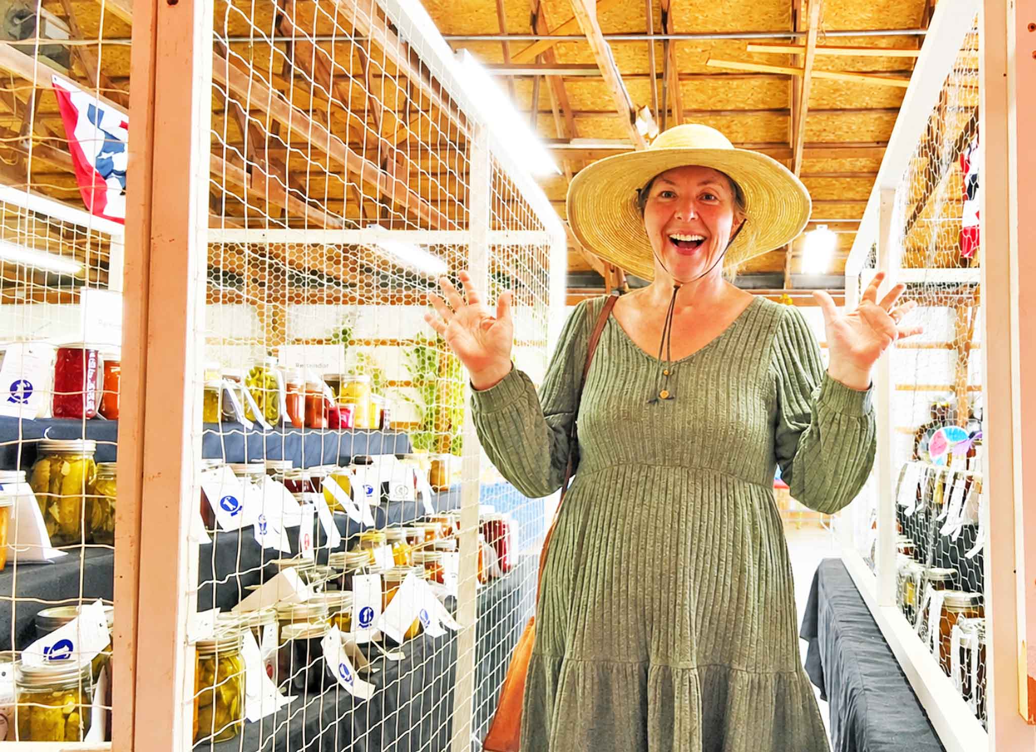 Jennifer with her hands raised in surprise beside shelves of ribboned canning jars at the Chelsea Community Fair.