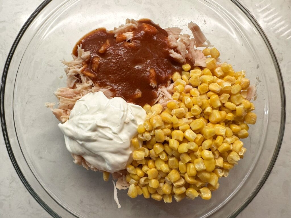 Sour cream, red enchilada sauce, and corn added to shredded chicken in a glass bowl