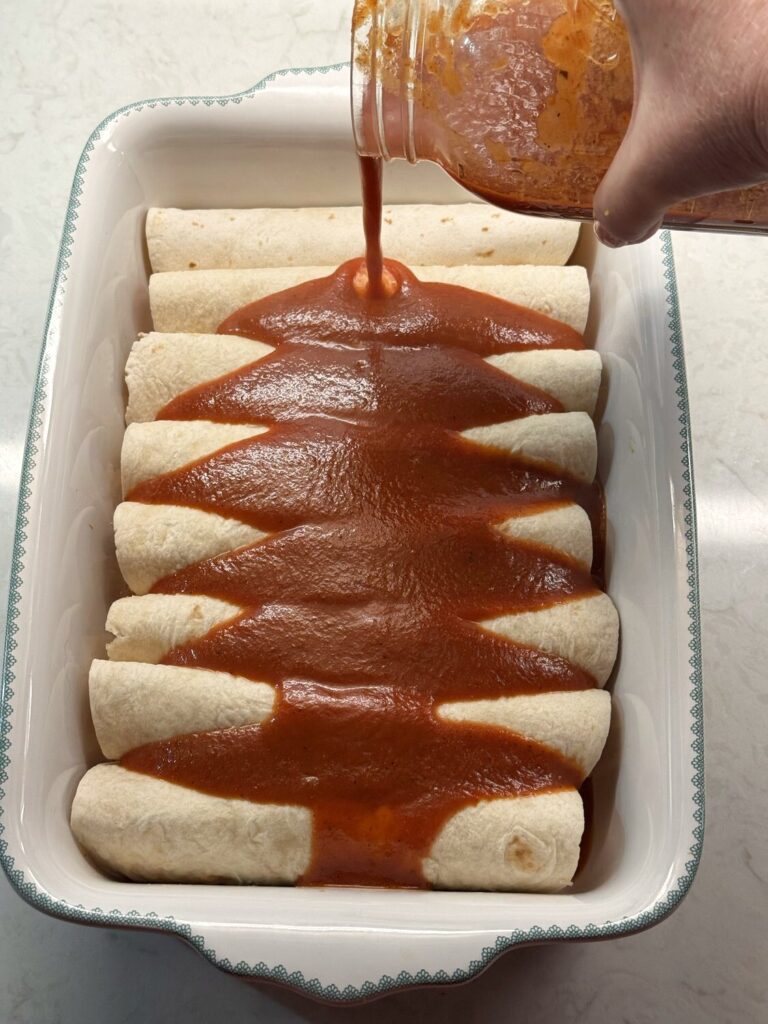 Red enchilada sauce being poured from a glass jar over rolled enchiladas