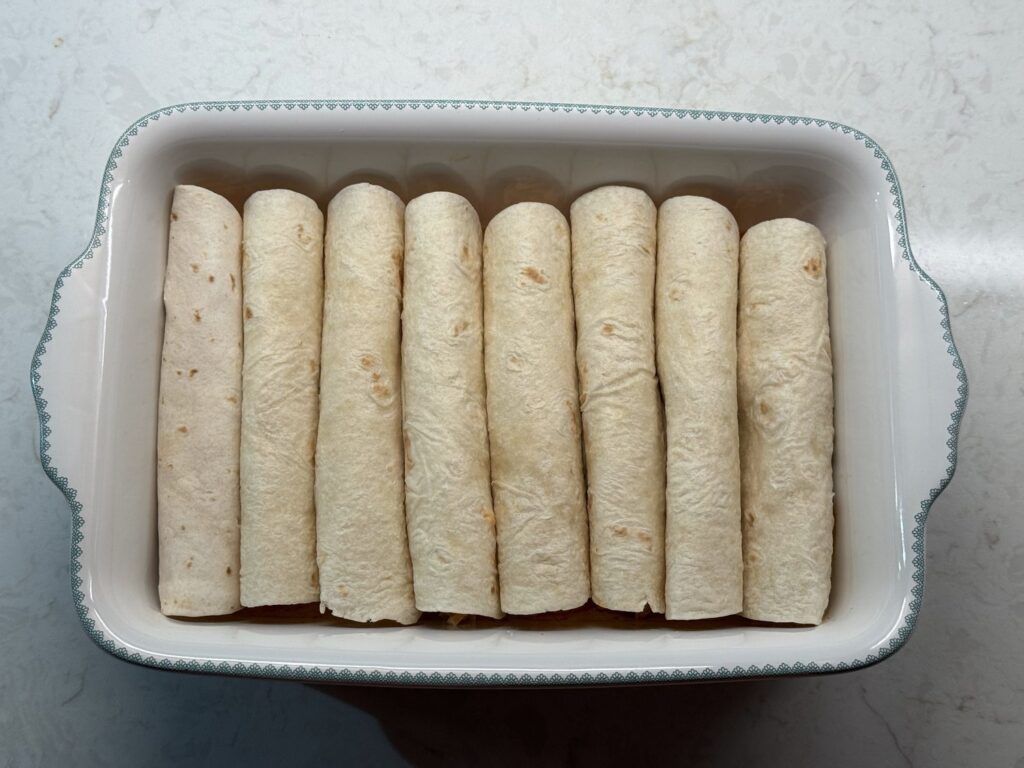 Eight rolled flour tortilla enchiladas lined up seam side down in a white baking dish