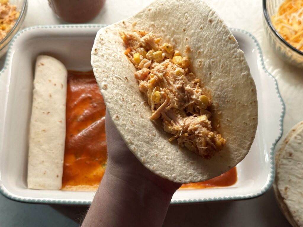 A flour tortilla held over the baking dish with a line of chicken, corn, and cheese filling down the middle